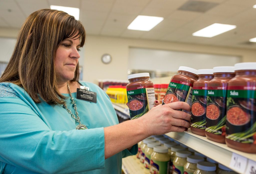 A senior service missionary wearing a missionary badge stocking canned food items on shelves inside a clean Bishop's Storehouse facility.